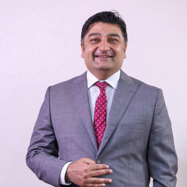 Rashid Khalani, CEO of Aga Khan University Hospital, in a grey suit with a red patterned tie, smiling confidently against a plain background. Portrait of Rashid Khalani, Chief Executive Officer of Aga Khan University Hospital, standing in a grey business suit, symbolising leadership in healthcare transformation.