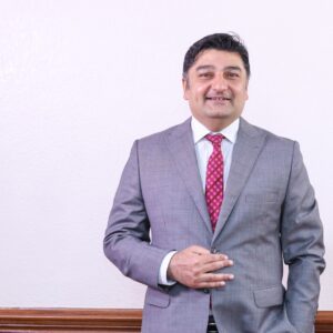 Rashid Khalani, CEO of Aga Khan University Hospital, in a grey suit with a red patterned tie, smiling confidently against a plain background. Portrait of Rashid Khalani, Chief Executive Officer of Aga Khan University Hospital, standing in a grey business suit, symbolising leadership in healthcare transformation.
