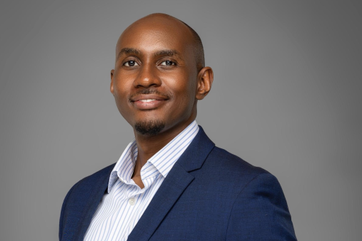 Kenneth Muhangi, a distinguished Ugandan lawyer, lecturer, and reform advocate, is pictured standing confidently in a professional setting, dressed in a navy-blue suit and a light-striped shirt. He is a Partner at KTA Advocates, a leading law firm specializing in technology, intellectual property, sustainability, and policy law. Known for his calm, thoughtful demeanor and sharp intellect, Kenneth embodies a new generation of legal leadership rooted in integrity, practical service, and ethical reform. His career reflects a commitment to mentorship, innovation, and institutional strengthening, while his personal philosophy balances impact, mindfulness, and faith-based purpose. Through his work in Uganda and across East Africa, he continues to champion technology-driven legal reform, education, and empowerment of young lawyers, promoting a legal profession defined by discipline, collaboration, and social responsibility.