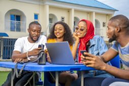 Four students sitting outdoors around a laptop and smartphones, discussing and reviewing study abroad options together.